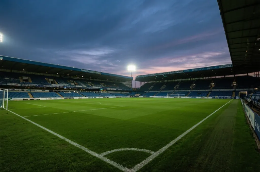 A wide-angle shot of a generic football stadium at dusk, empty lush green pitch, dramatic sky, cinematic sports photography